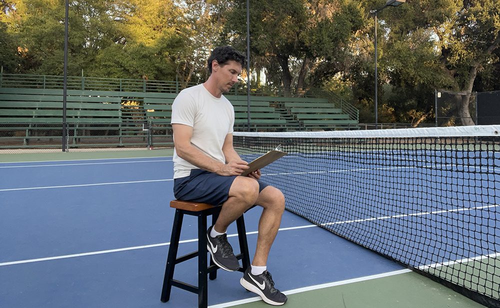 Jeremy sits courtside at a blue and green tennis court, looking over notes on a clipboard